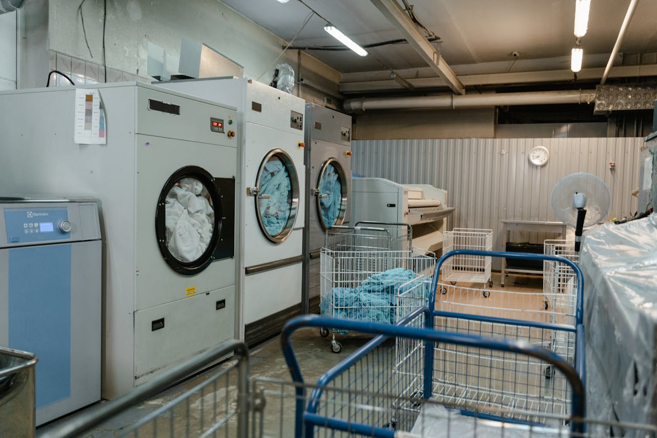hero-img-02 Interior view of an industrial laundry room with washers and rolling carts.